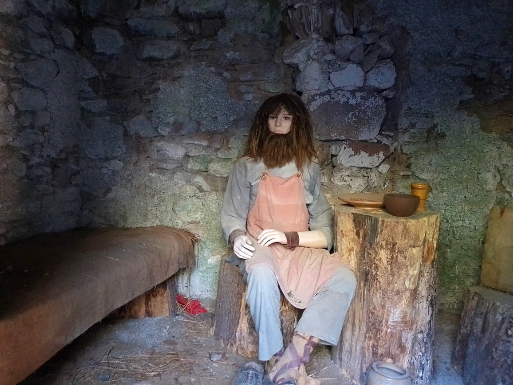 Gefangeneer in einem kleine Verließ - Puppe mit langem Bart, roter Schürze und hellblauer Hose, daneben eine Pritsche und ein Tisch mit Teller und Schüssel