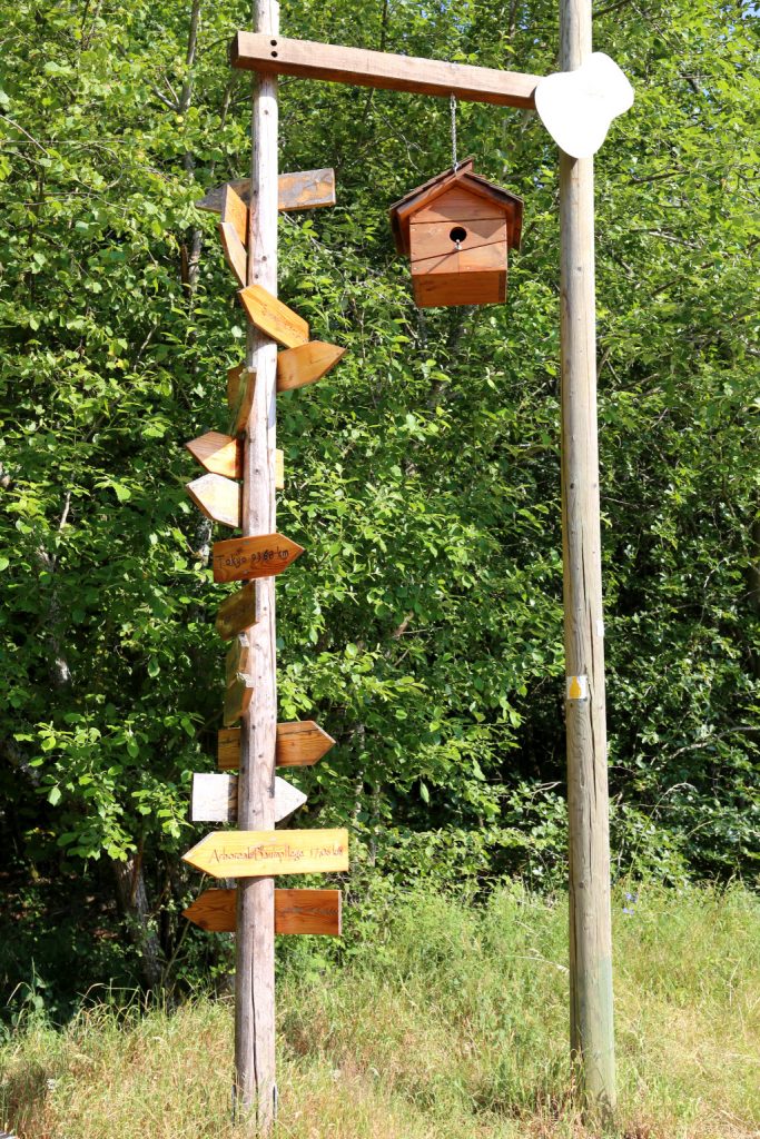 Wegweiser mit vielen kleinen Tafeln, die in alle Welt zeigen und einem Vogelhaus, alles aus Holz