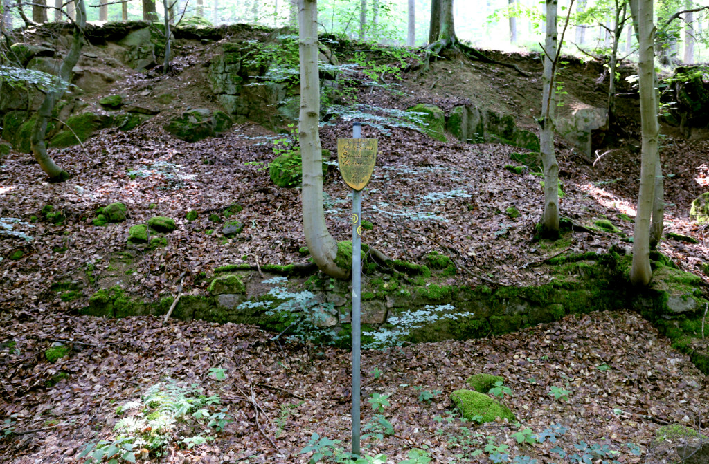 Wappenförmiges altes Schild auf Metallpfosten vor verfallenen Mauern im Wald