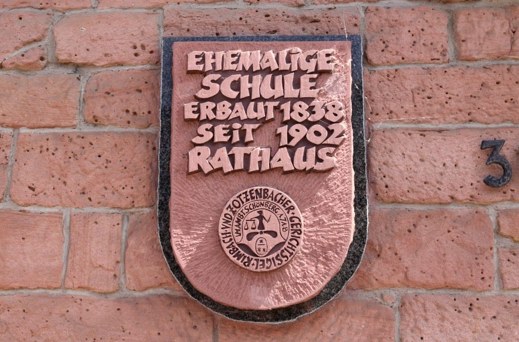 Schild aus rotem Sandstein mit Aufschrift: Ehemalige Schule, erbaut 1838, seit 1902 Rathaus; darunter Siegel des Rimbacher und Zotzenbacher Gerichtsstandes im Amt Schöberg 1718